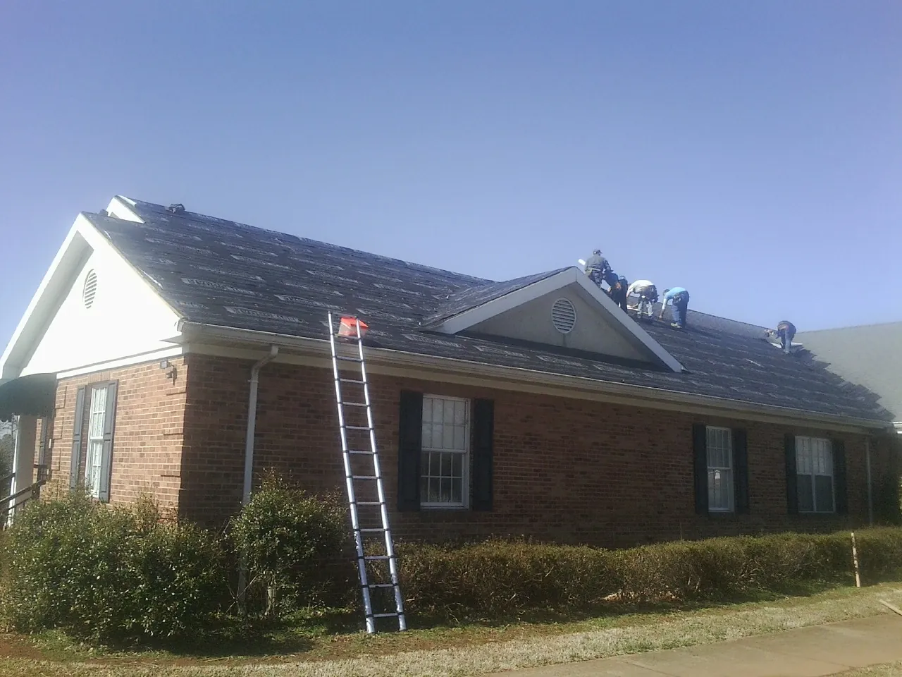 Crew tearing shingles off a brick ranch roof with a ladder set — Metal Roof Repair work by our crew in Lake Preston