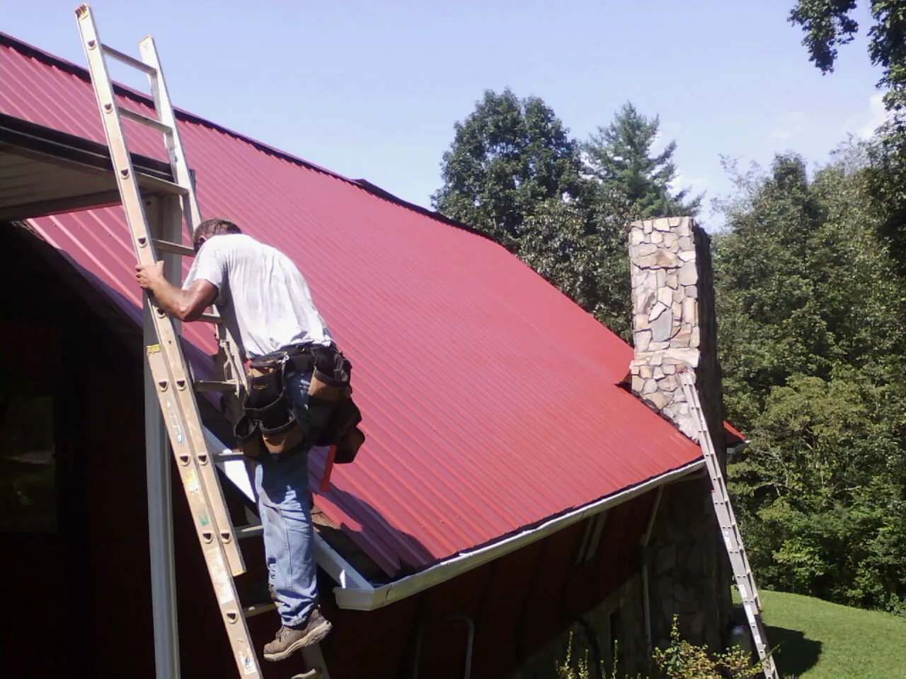 Roofer on a ladder beside a red metal roof and stone chimney — Soffit & Fascia Repair by Roofing Brothers in Lake Preston